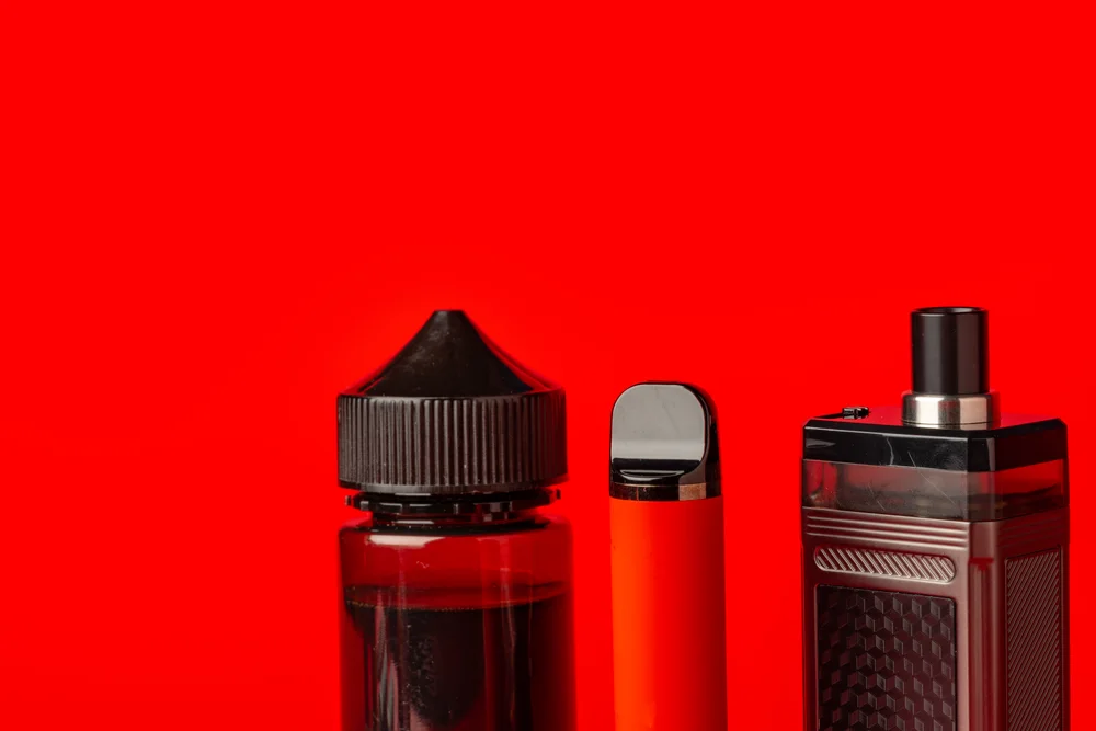 Liquidflasche, Pod-Gerät und E-Zigarette nebeneinander vor rotem Hintergrund